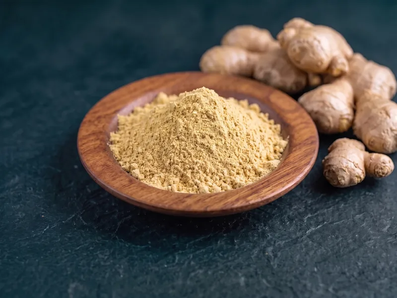 Dehydrated ginger powder in a wooden dish beside whole ginger rhizome